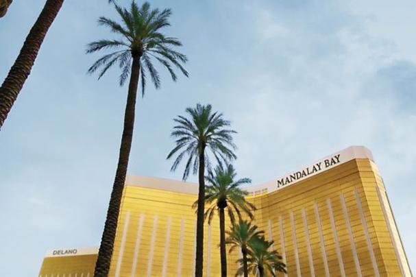 Tall palm trees stand in front of the gold Mandalay Bay hotel, viewed from below against a light blue sky.