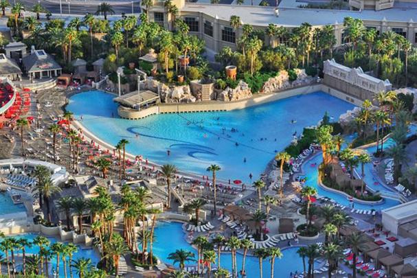 A wide aerial view of a large resort pool area with a wave pool, palm trees, and people swimming and lounging.